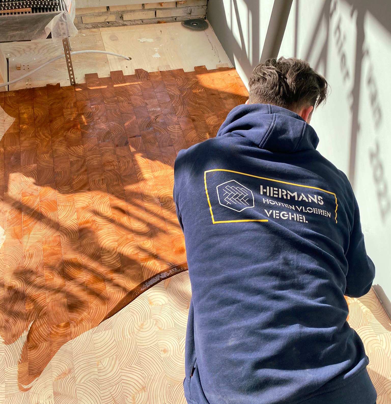 Ronald Hermans aan het werk aan een kopshouten vloer, in een trui met het logo van Hermans Houten Vloeren